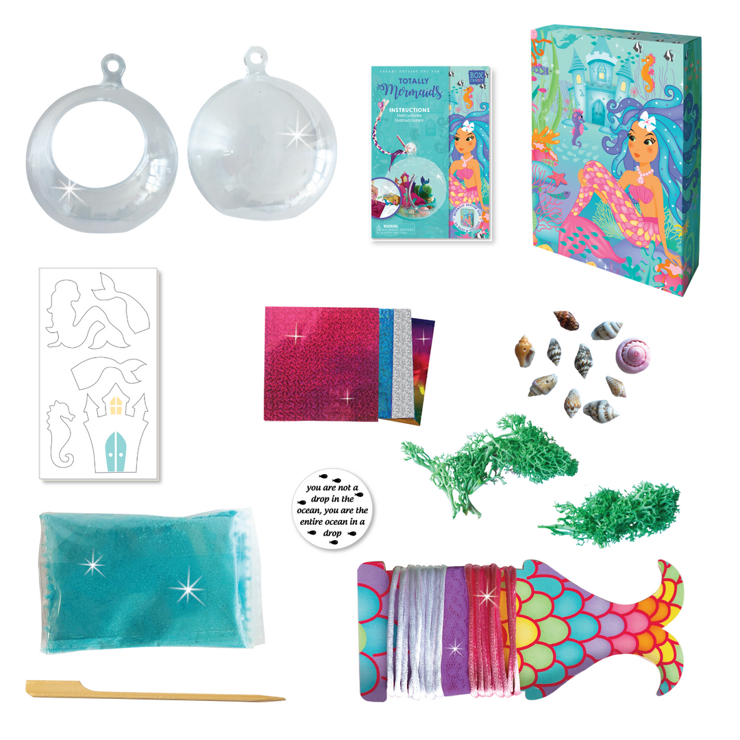 Out of box image of the materials that come with the Mermaid Terrarium which Includes: 1 plastic terrarium bowl, play foam "ocean", 4 cardboard figures with layer of sticky paper, 4 foil sheets, 1 wooden craft stick, green decorative seaweed plants, approximately 10 decorative shells, stickers, colored lace, strings and instructions, all in a beautiful keepsake storage box made from recycled cardboard.