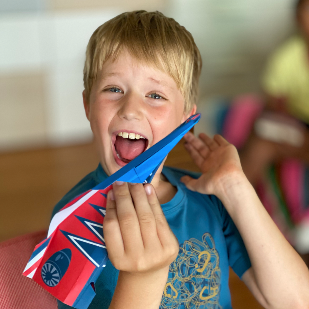 Lifestyle image of a child smiling showing his finished paper airplane. 